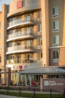 Un hotel de varios pisos con un diseño moderno, con balcones en cada planta y un restaurante visible llamado "CRAVE" a nivel de calle.