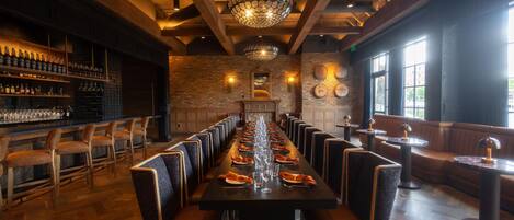 A long dining table with chairs, a bar area with bottles, and a brick wall with a chandelier.