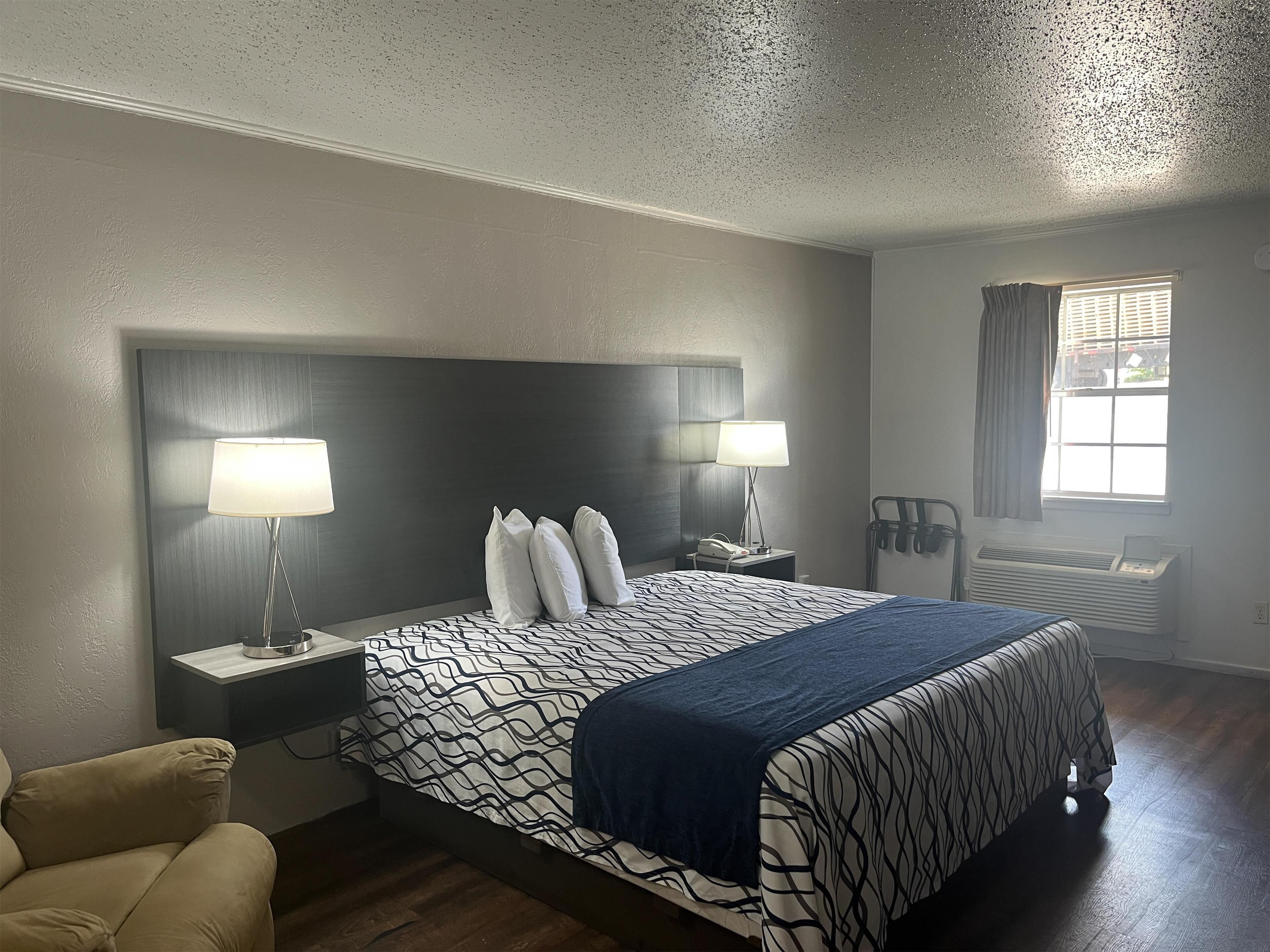 A hotel room with a large bed, two bedside tables with lamps, a chair, a window with curtains, and a wall-mounted heater.