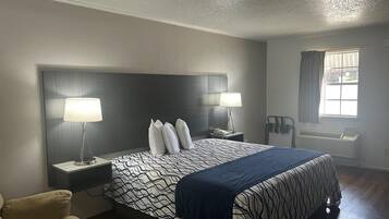 A hotel room with a large bed, two bedside tables with lamps, a chair, a window with curtains, and a wall-mounted heater.