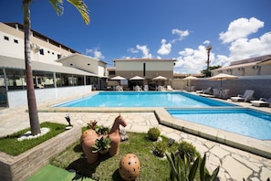 Piscina con sillas de descanso y sombrillas, rodeada por un jardín ajardinado con macetas decorativas y una palmera.