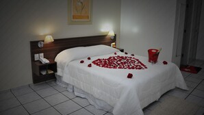 Un llit ben fet amb roba de llit blanca, un capçal de fusta, lampades laterals, un telèfon al costat del llit i un arranjament de roses vermelles damunt el llit.