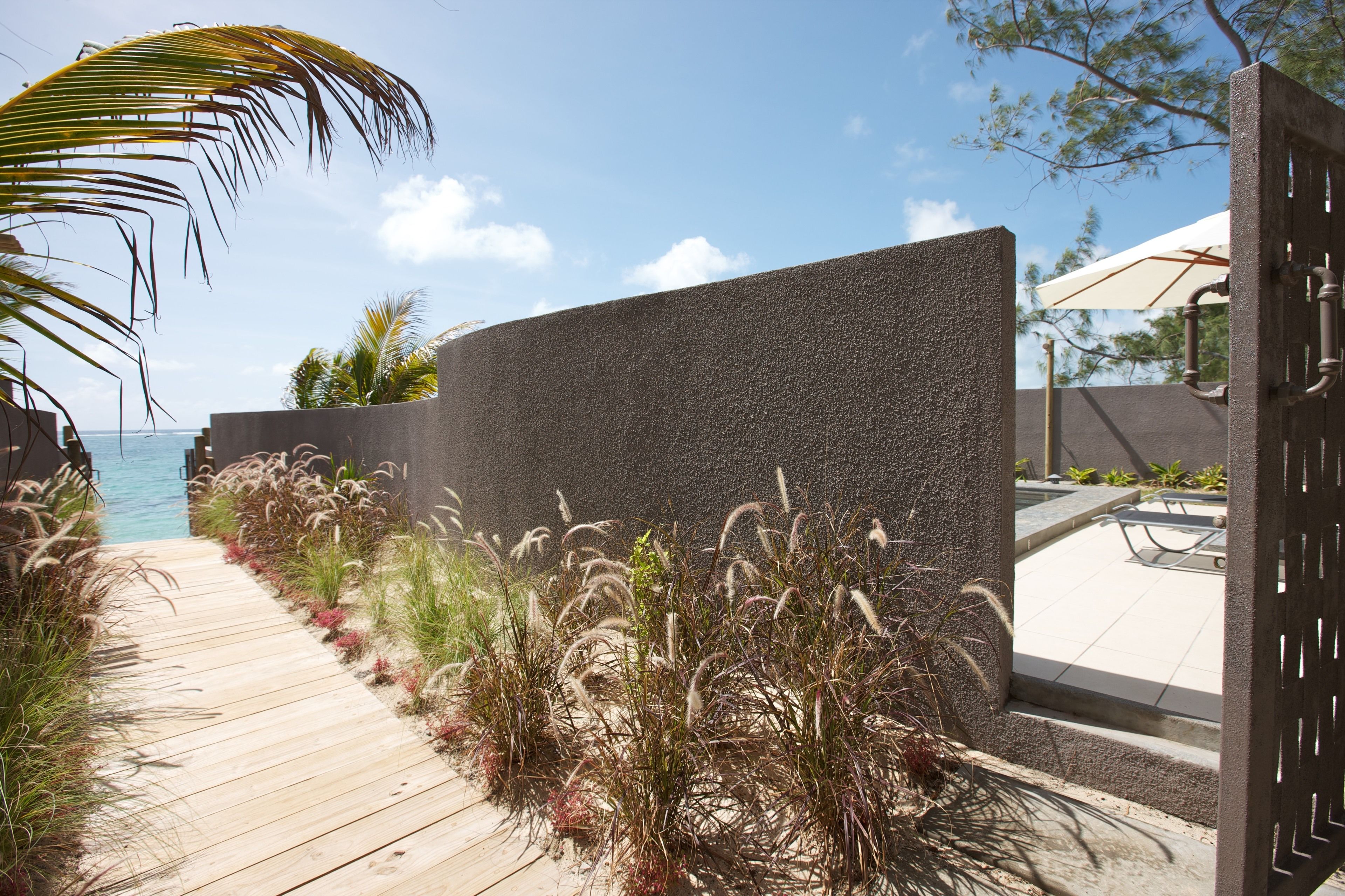 Un sendero de madera conduce a un edificio moderno con pared texturizada, rodeado de plantas tropicales y vista al océano.