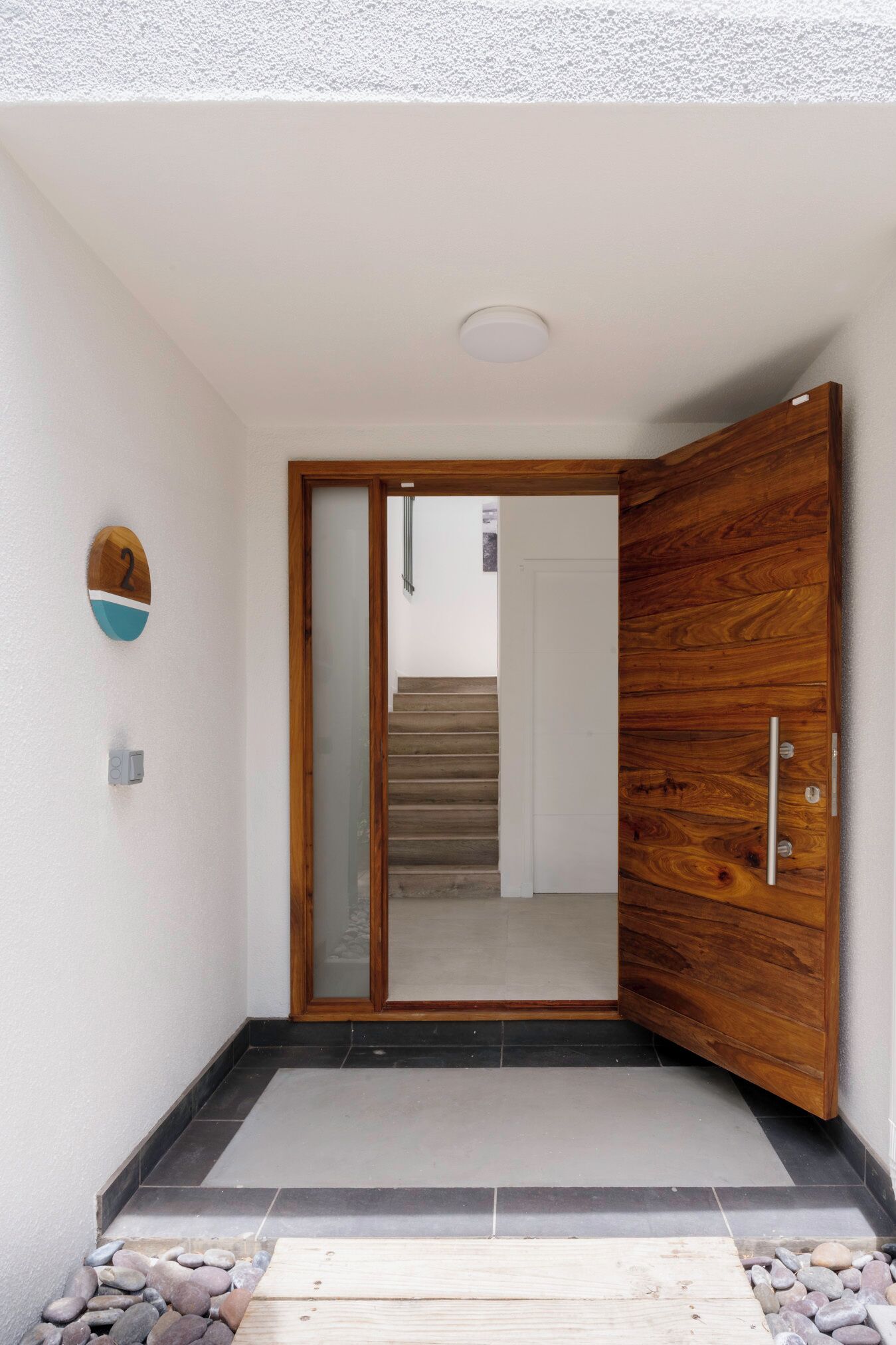 A modern entrance with a wooden door, a number plaque, and a stone pathway leading to a staircase.