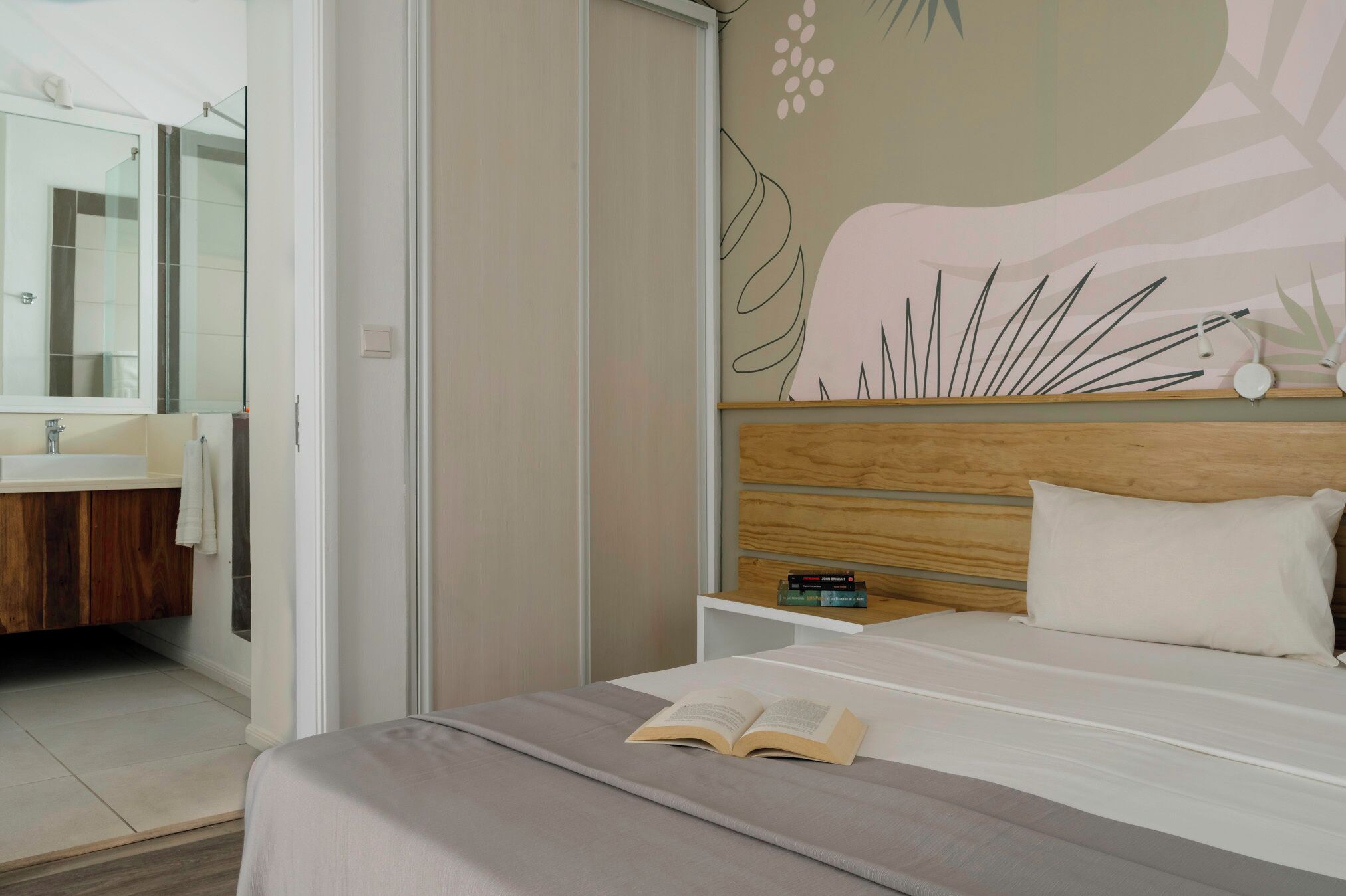Ein modernes Schlafzimmer mit einem großen Bett, einem Nachttisch mit Büchern, einem Holz-Kopfteil und einem Badezimmer mit Spiegel und Waschbecken.