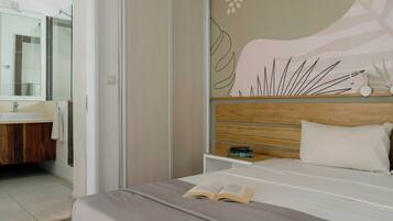 A modern bedroom with a large bed, a bedside table with books, a wooden headboard, and a bathroom with a mirror and sink.