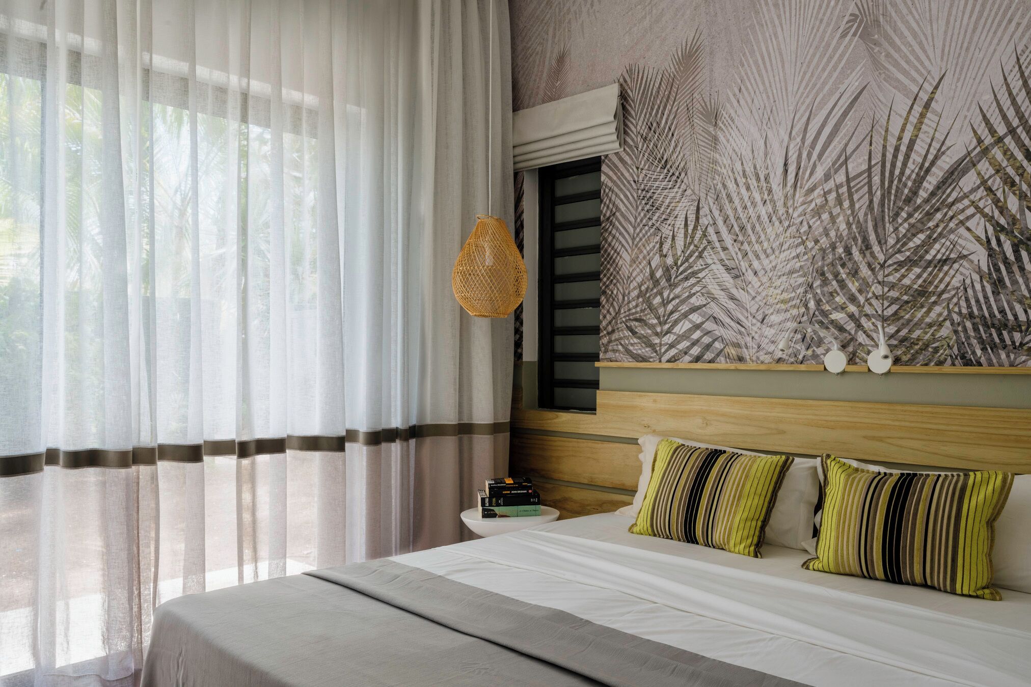 A neatly made bed with pillows, a bedside table with books, sheer curtains, and a wall with a leaf pattern.
