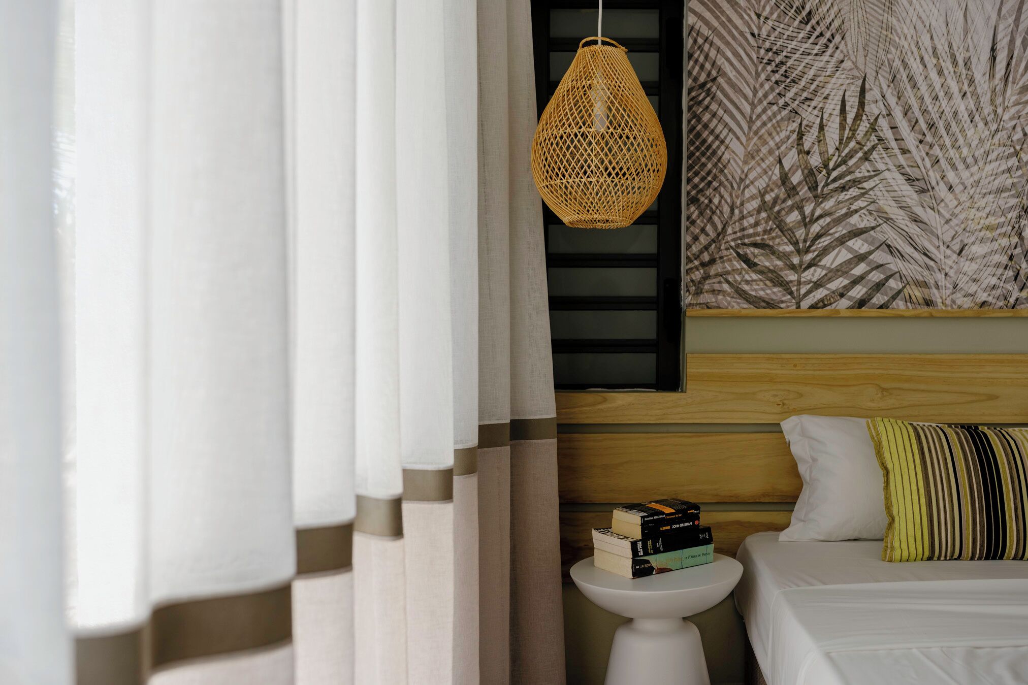 A bedroom with a bed, a bedside table with books, a hanging wicker lamp, and a wall with a leaf pattern.