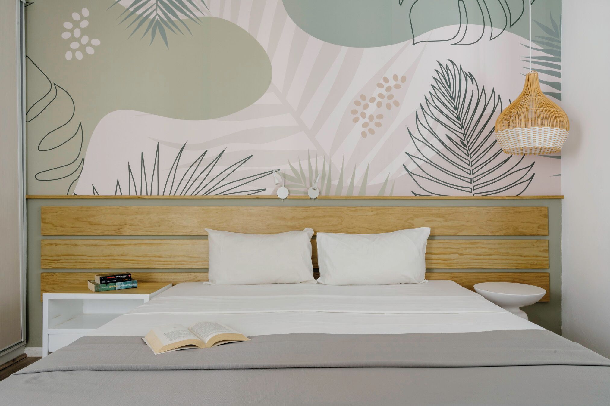 Un dormitorio con una cama, una mesita de noche con libros, una lámpara colgante y una pared con diseño de hojas.