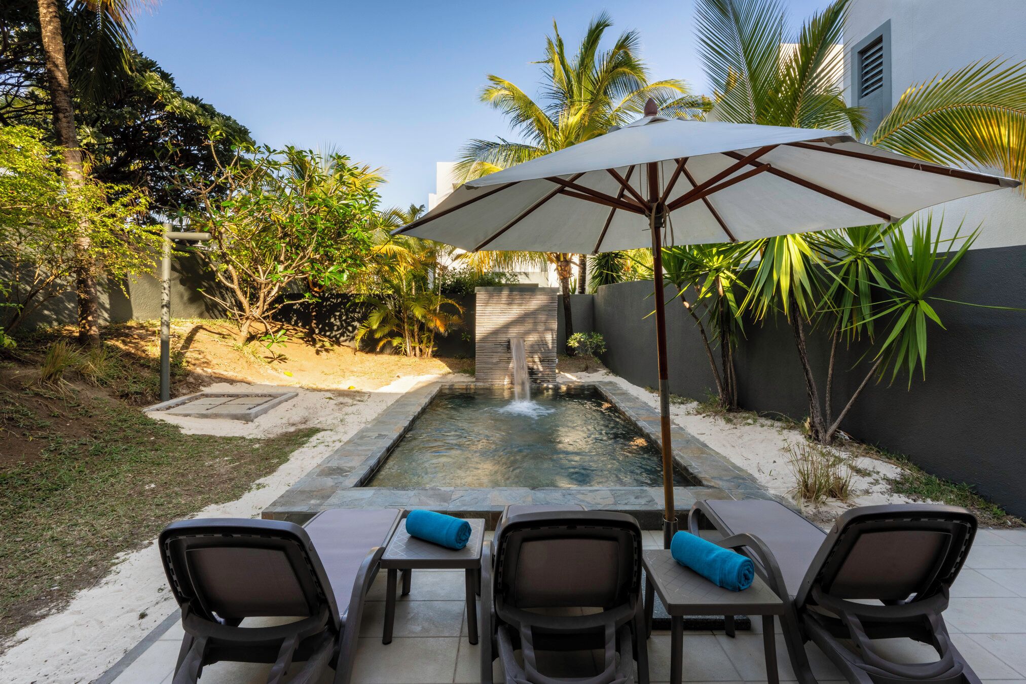Zona de piscina con una cascada, rodeada de palmeras y un área de estar cubierta con sillas y una sombrilla.