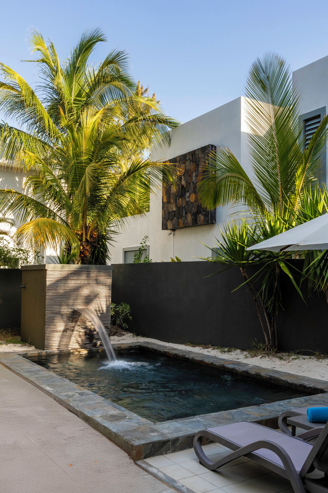 A swimming pool with a waterfall, surrounded by palm trees and a modern building with a stone wall.