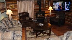 L’intérieur d’un chalet confortable, avec des murs en bois, une cheminée, deux fauteuils en cuir, un canapé et un téléviseur diffusant un programme d’information.