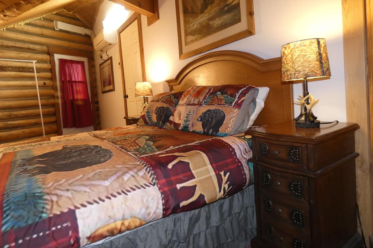 Une chambre confortable, aménagée dans le style d’un chalet en rondins, avec un grand lit, une tête de lit en bois, une lampe et une commode.