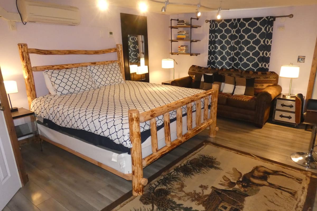 Un lit en bois avec une couvre-lit à motifs, une table de chevet en bois, une lampe de chevet, un tapis à motifs, un canapé en cuir marron et une étagère murale avec des livres.