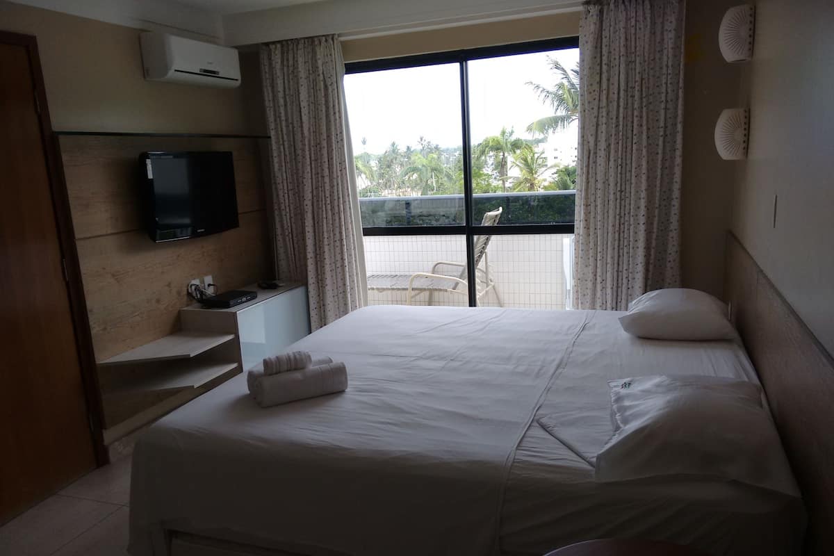 Une chambre d’hôtel comprenant un lit, un téléviseur fixé au mur, un bureau avec une chaise et un balcon offrant une vue sur les palmiers et les immeubles.