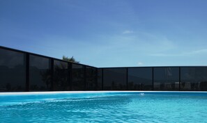 Un área de piscina moderna con agua azul cristalina, rodeada por un edificio de vidrio con vistas a palmeras y montañas al fondo.