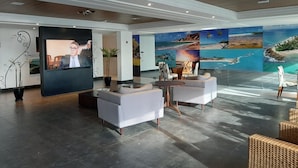 Een moderne lobby met een groot televisiescherm, comfortabele zitplaatsen en een muur met tropische strandmuurschilderingen.