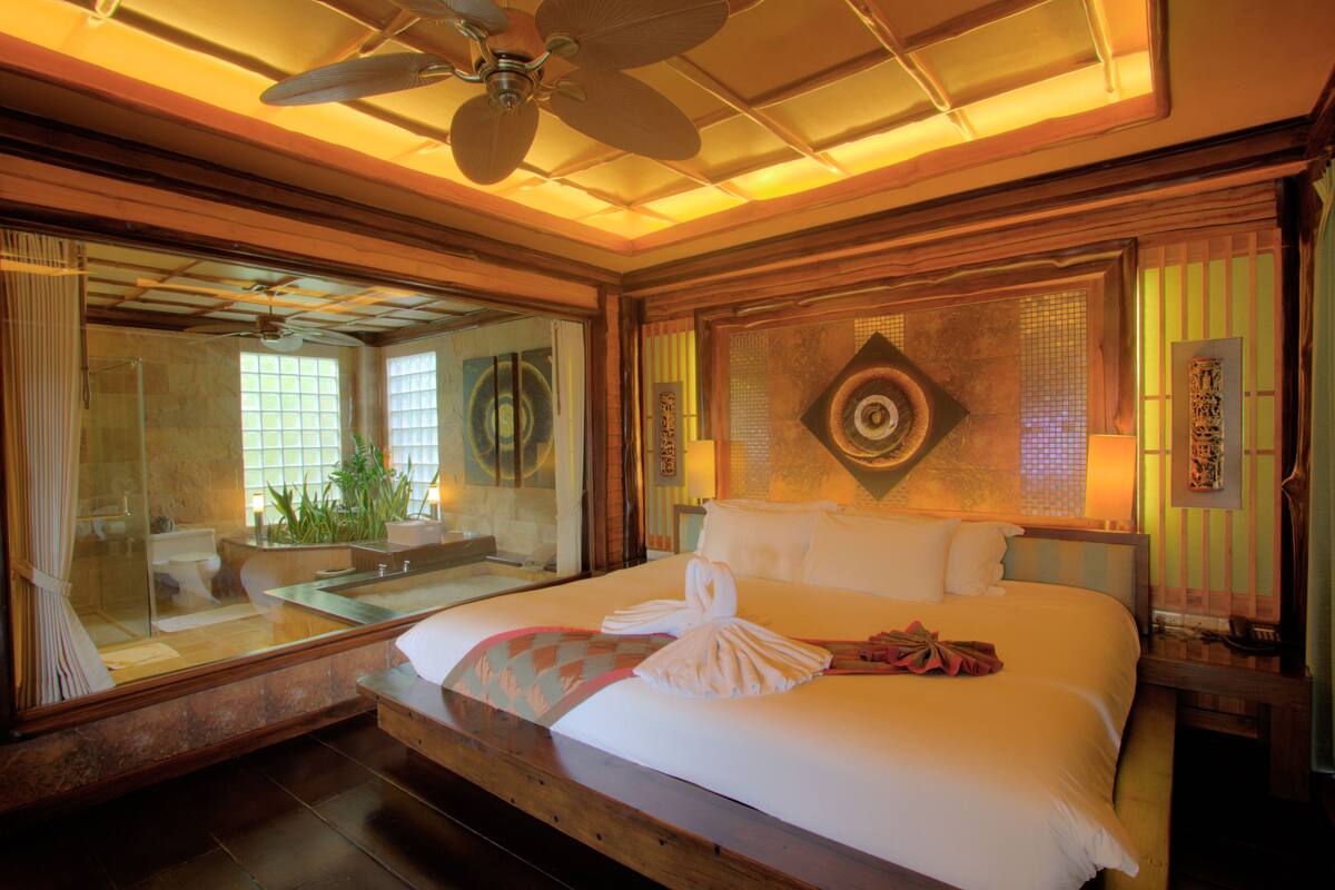 A spacious hotel room with a large bed, a ceiling fan, a glass shower enclosure, and a view of the bathroom.