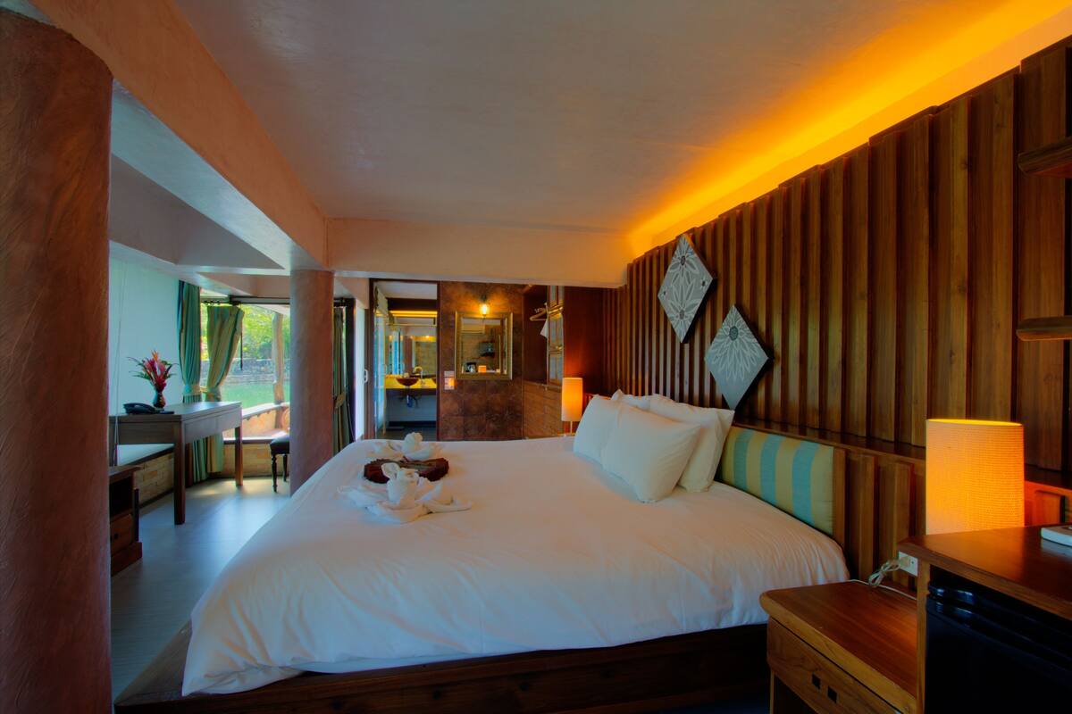 A spacious bedroom with a large bed, wooden paneling, a desk with a chair, and a view of a bathroom.