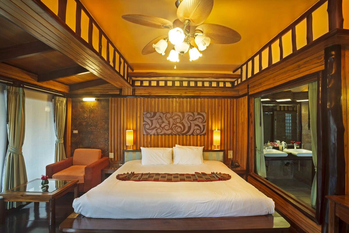 A hotel room with a large bed, a wooden ceiling fan, a sitting area with a chair, and a large window.