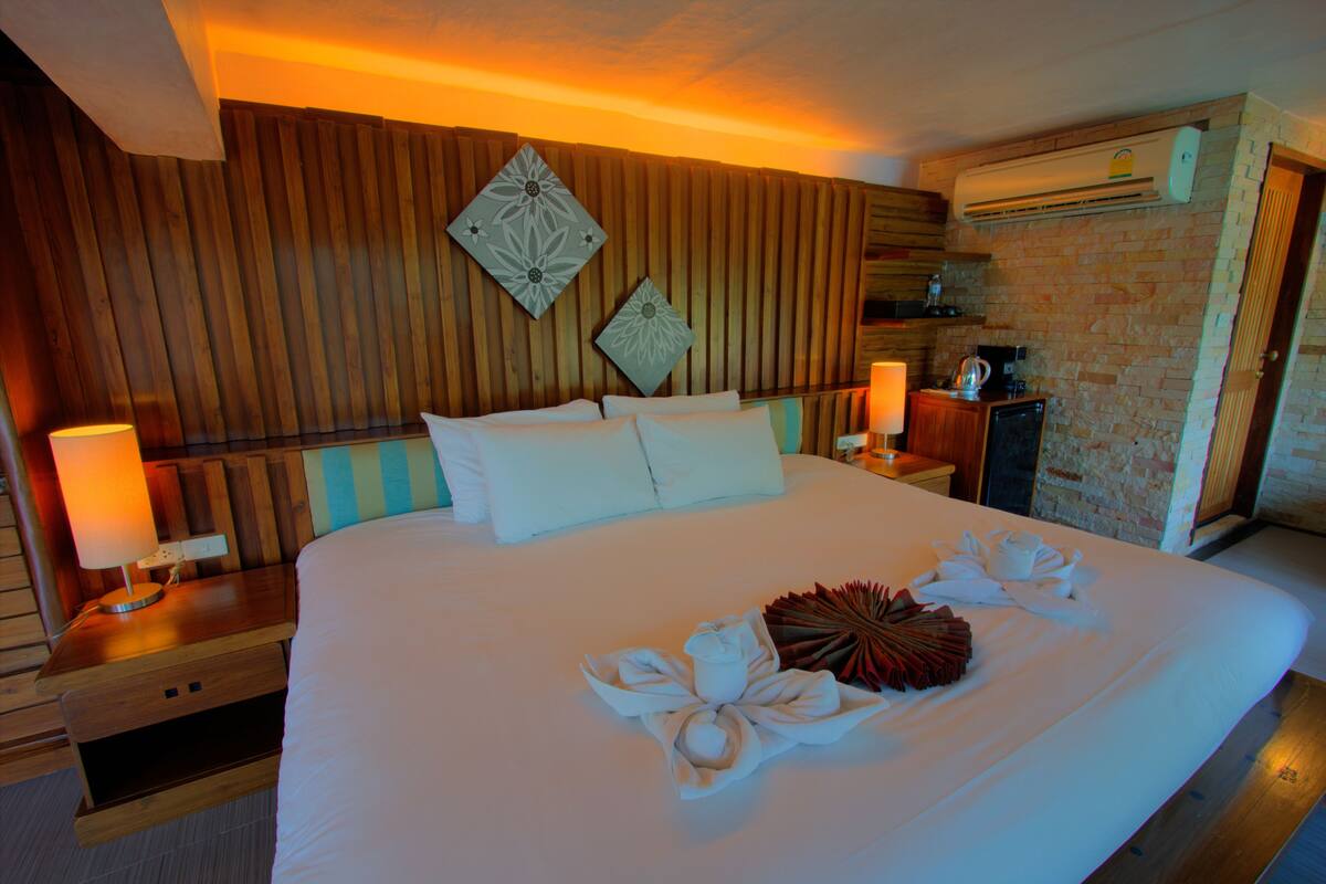 A hotel room with a large bed, wooden headboard, two wall-mounted decorative pillows, a bedside table with a lamp, and an air conditioning unit.