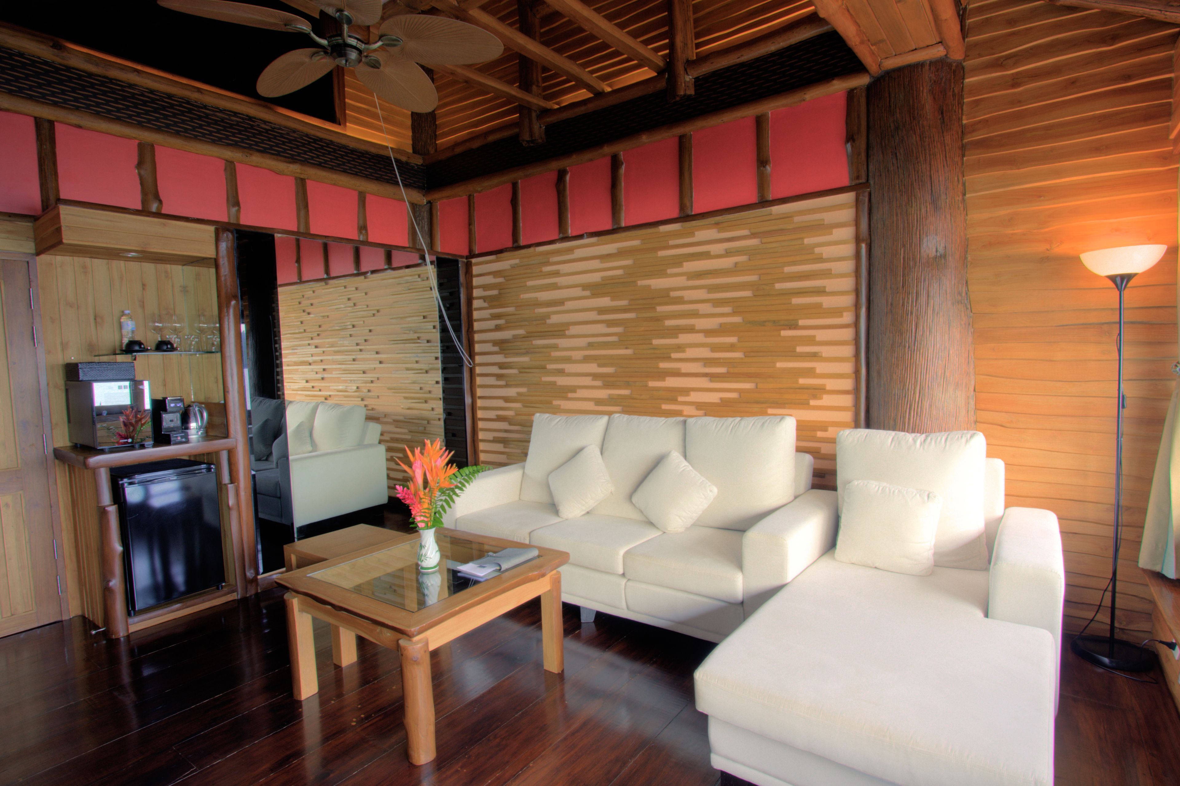 A modern living room with a wooden ceiling, a white sofa, a small table with a vase of flowers, and a compact kitchen area in the background.