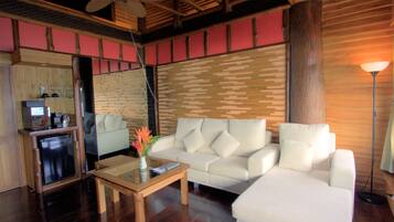 A modern living room with a wooden ceiling, a white sofa, a small table with a vase of flowers, and a compact kitchen area in the background.