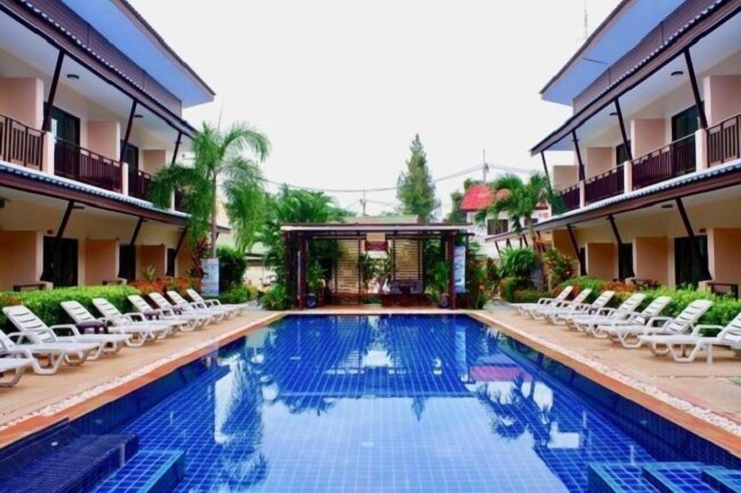 Una piscina con sillas de descanso y una zona de estar cubierta, rodeada de vegetación y edificios de dos pisos.