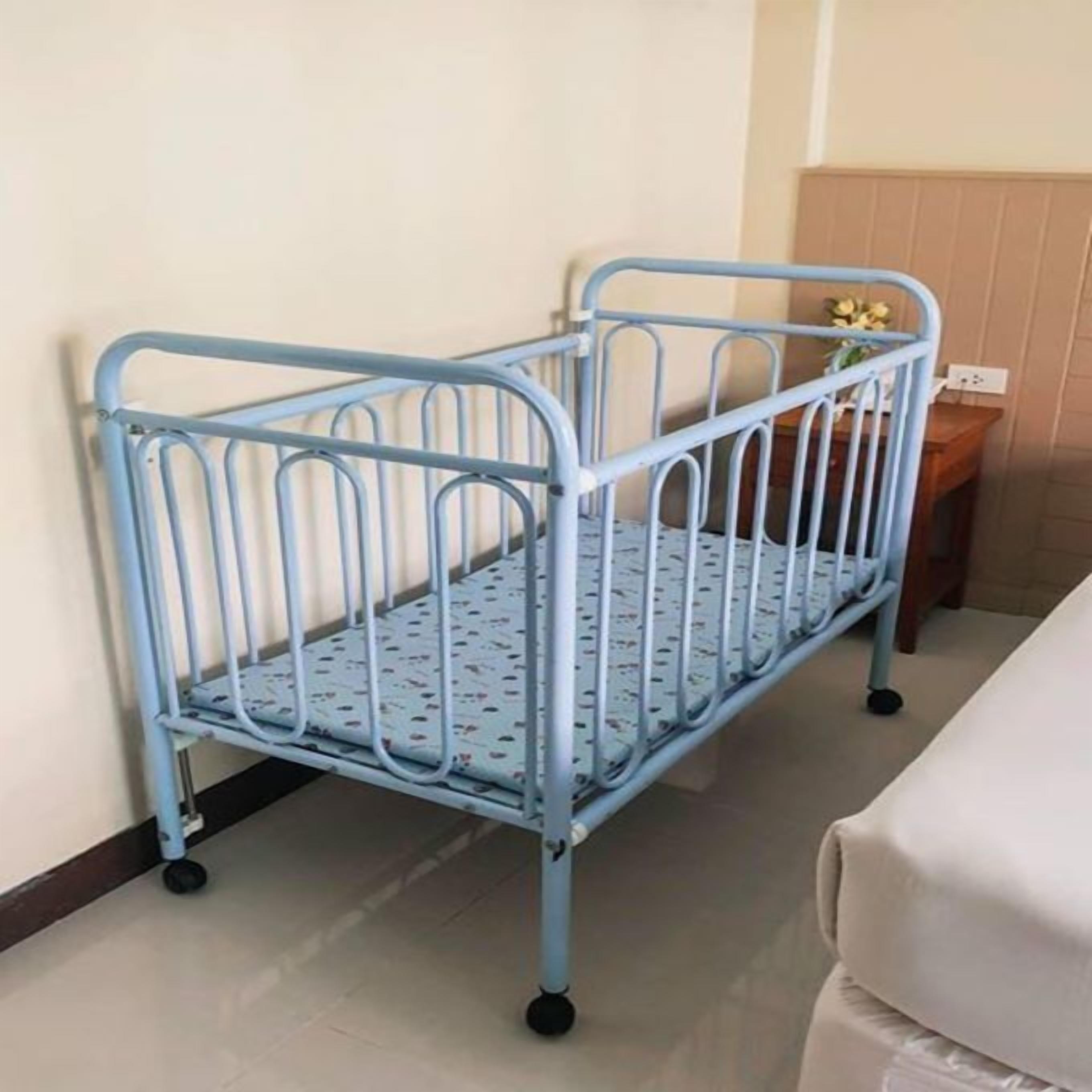 A metal crib with wheels, positioned against a wall in a room with a wooden floor.