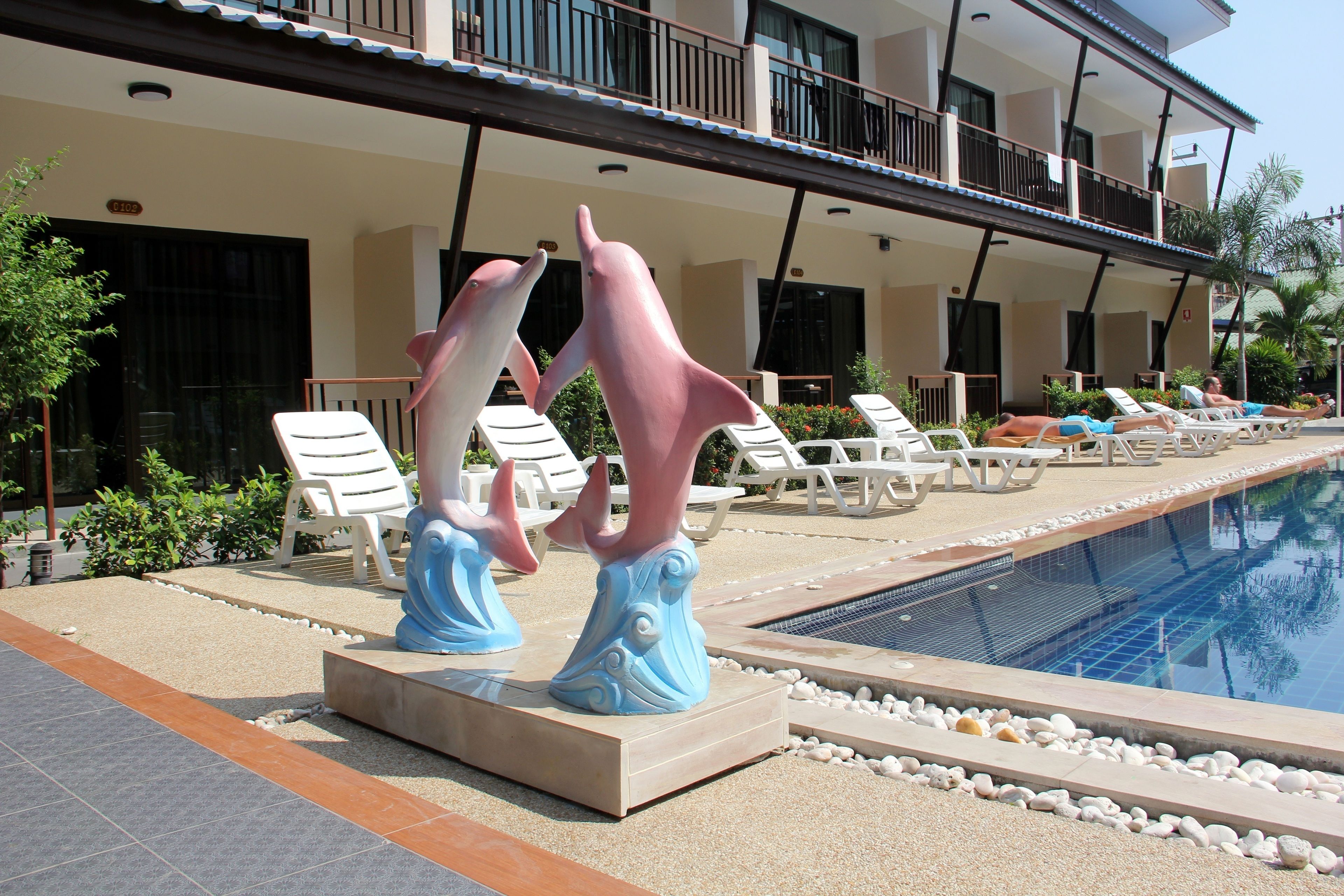A pool area with lounge chairs, a decorative dolphin sculpture, and a building with balconies.