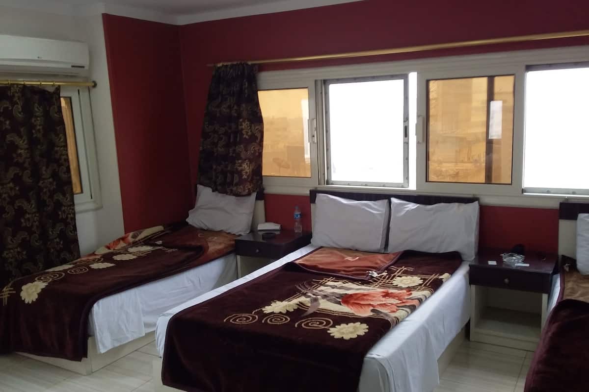A hotel room with two beds, a window with curtains, a bedside table with a lamp, and a wall-mounted air conditioner.