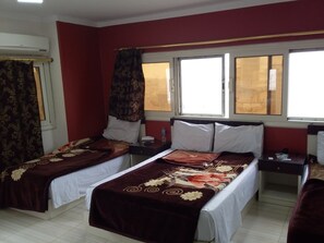 A hotel room with two beds, a window with curtains, a bedside table with a lamp, and a wall-mounted air conditioner.