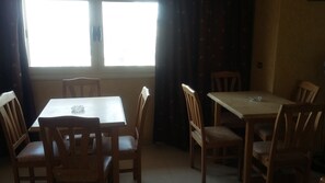 A dining area with two wooden tables and chairs, a window with curtains, and a tiled floor.