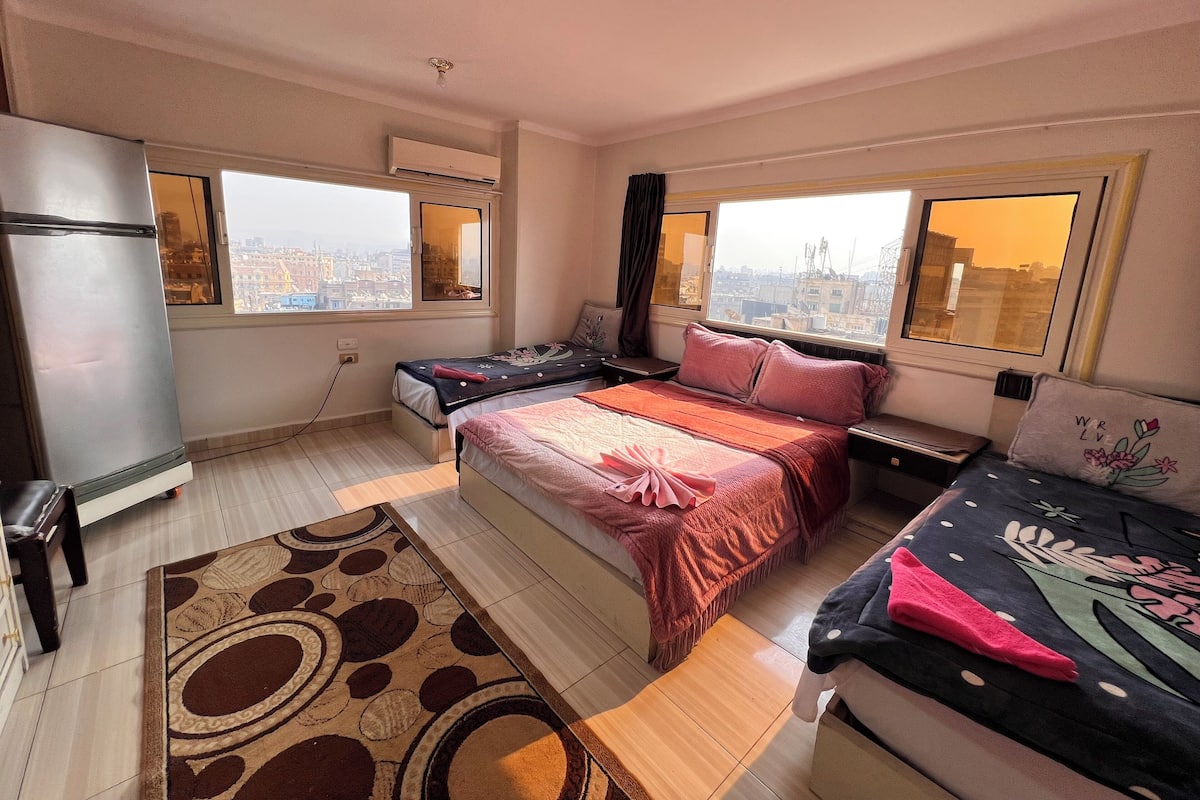 A bedroom with two beds, a refrigerator, and a window with a city view.