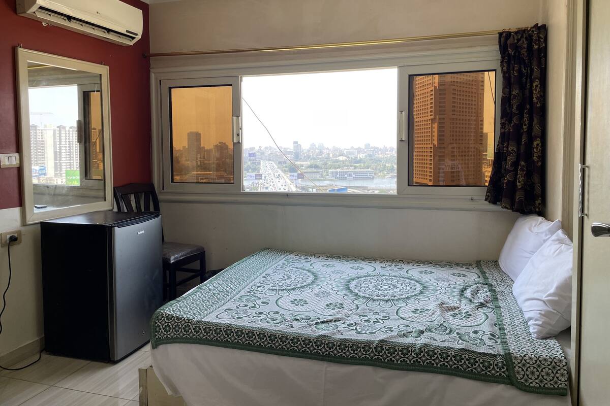 A bedroom with a bed, a desk, an air conditioner, and a window with a city view.