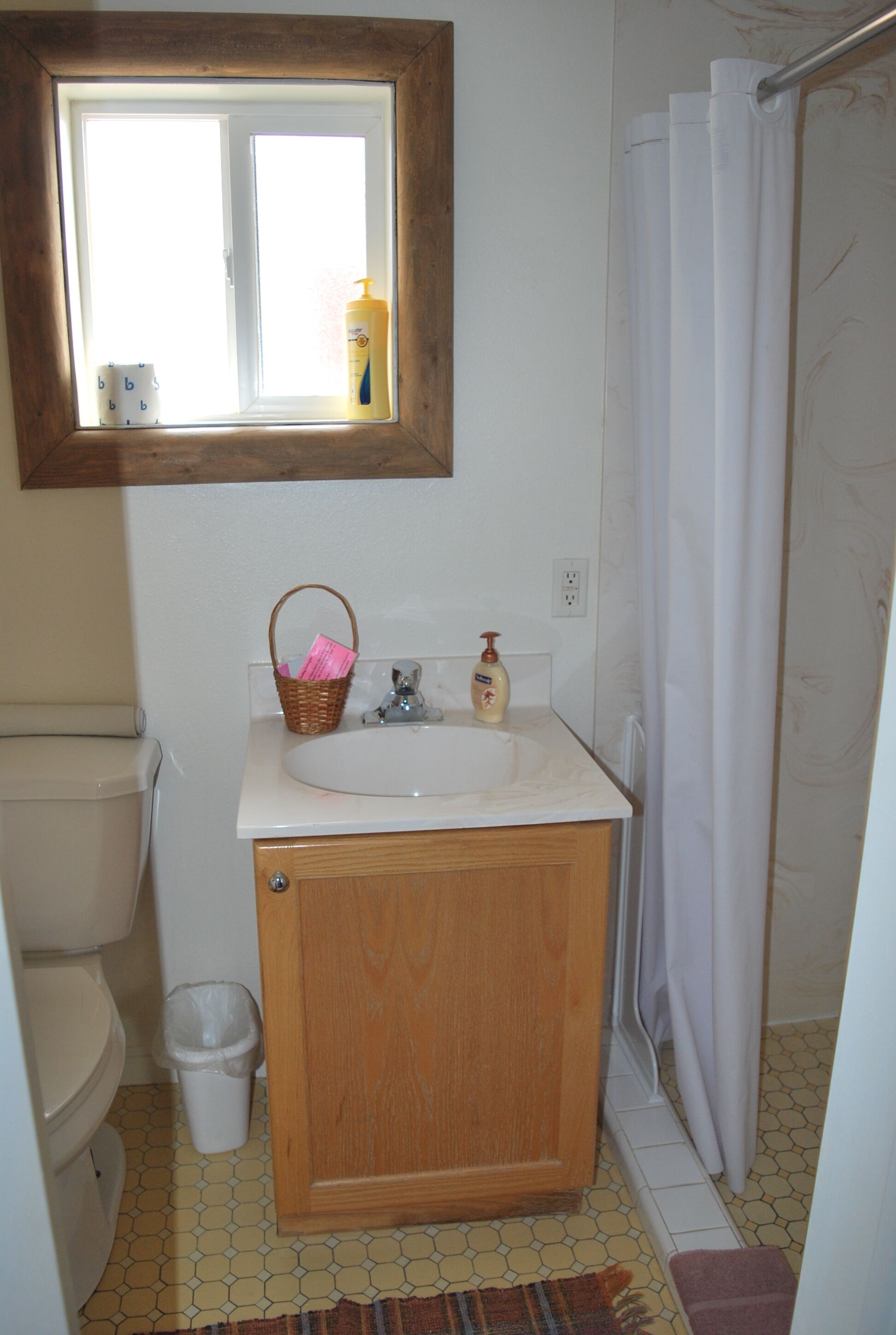 Un baño con un mueble de madera, un lavabo, una ventana, una cortina de ducha y un cubo de basura.
