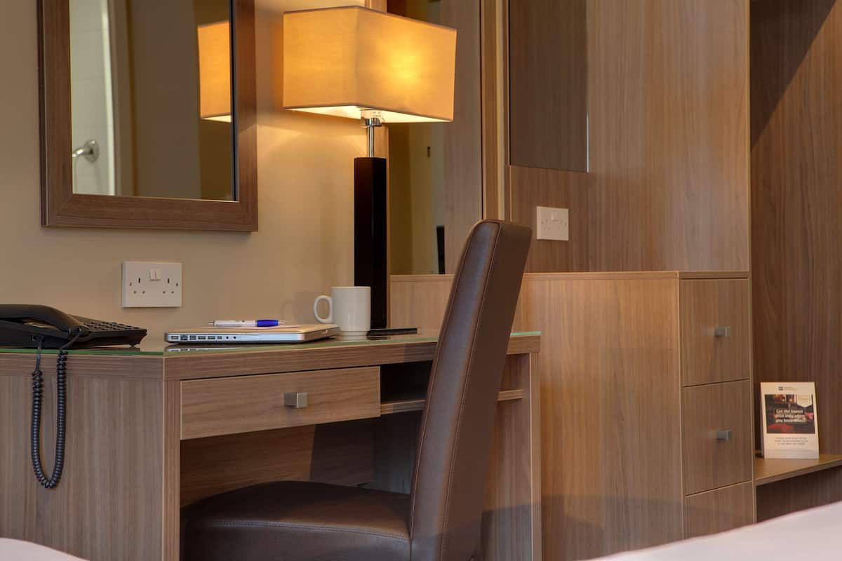 A hotel room with a wooden desk, a chair, a lamp, a phone, a laptop, a mug, and a mirror.