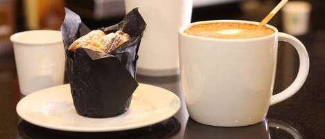A cup of coffee with a spoon, a pastry in a paper wrapper, and a white cup on a table.