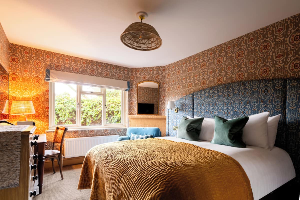 A bedroom with a large bed, a desk, a chair, a window, and patterned wallpaper.