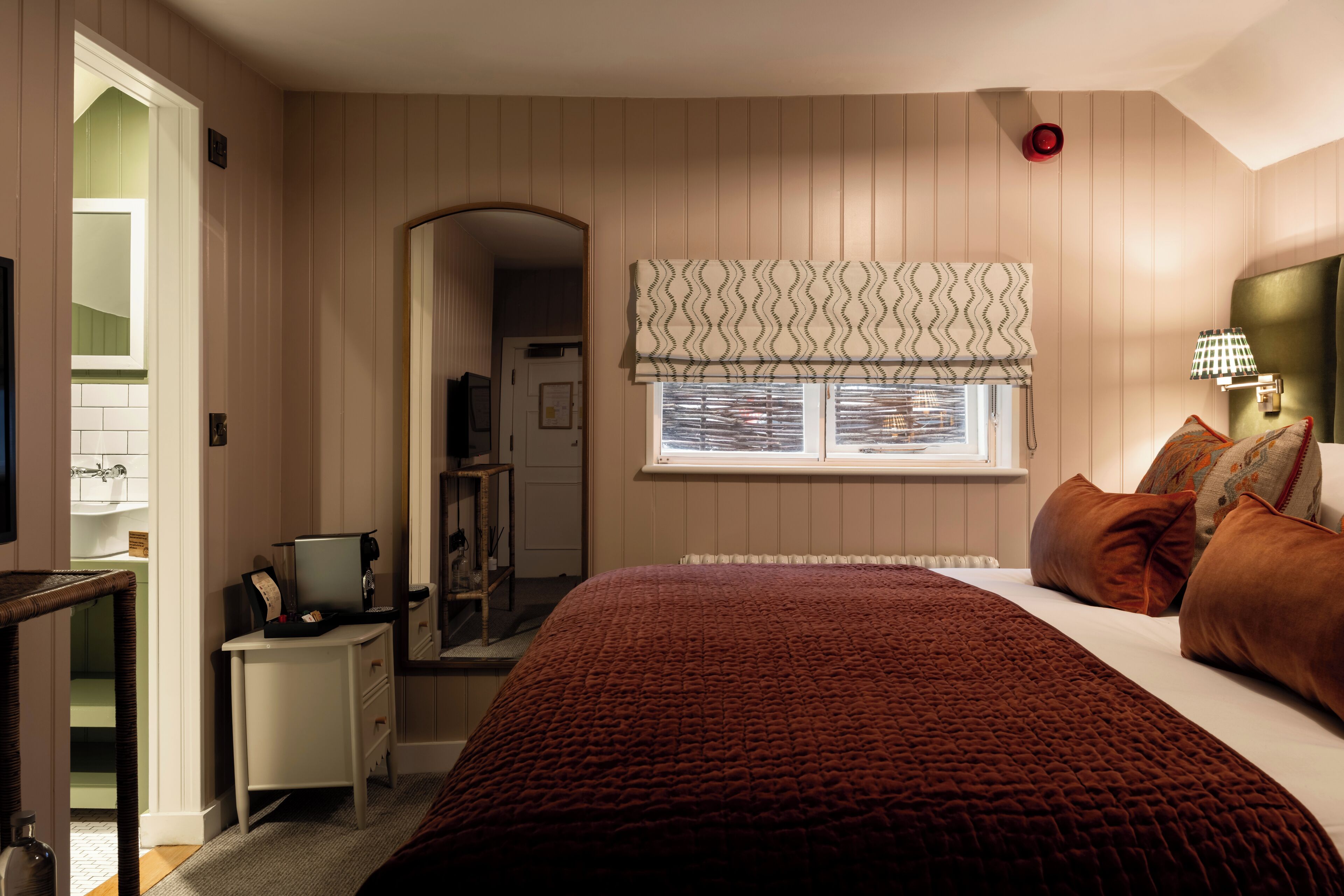 A bedroom with a bed, bedside table, lamp, window with blinds, and a door leading to a bathroom.