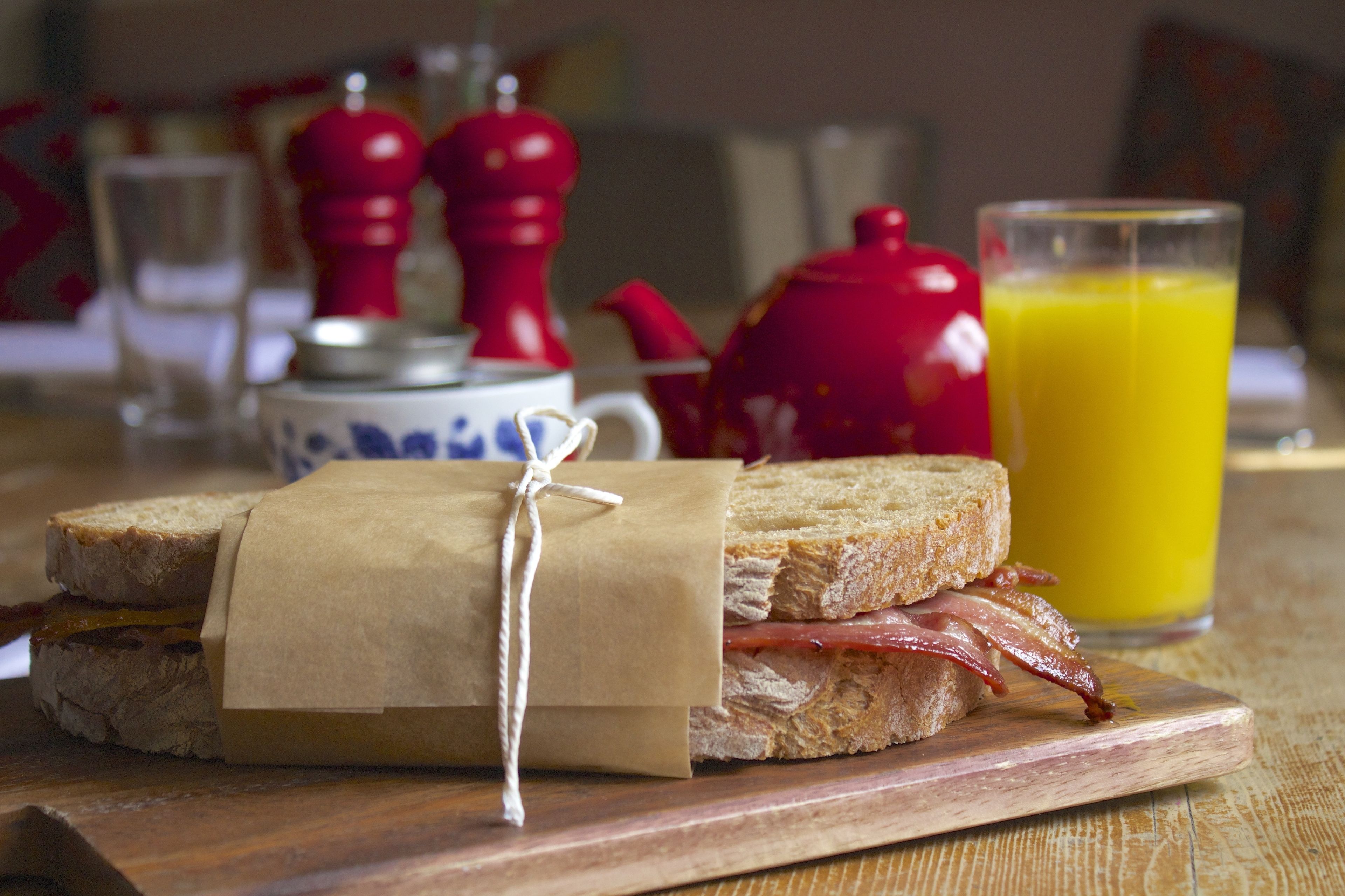 Un sándwich de desayuno sobre una tabla de madera, un vaso de jugo de naranja y una tetera al fondo.