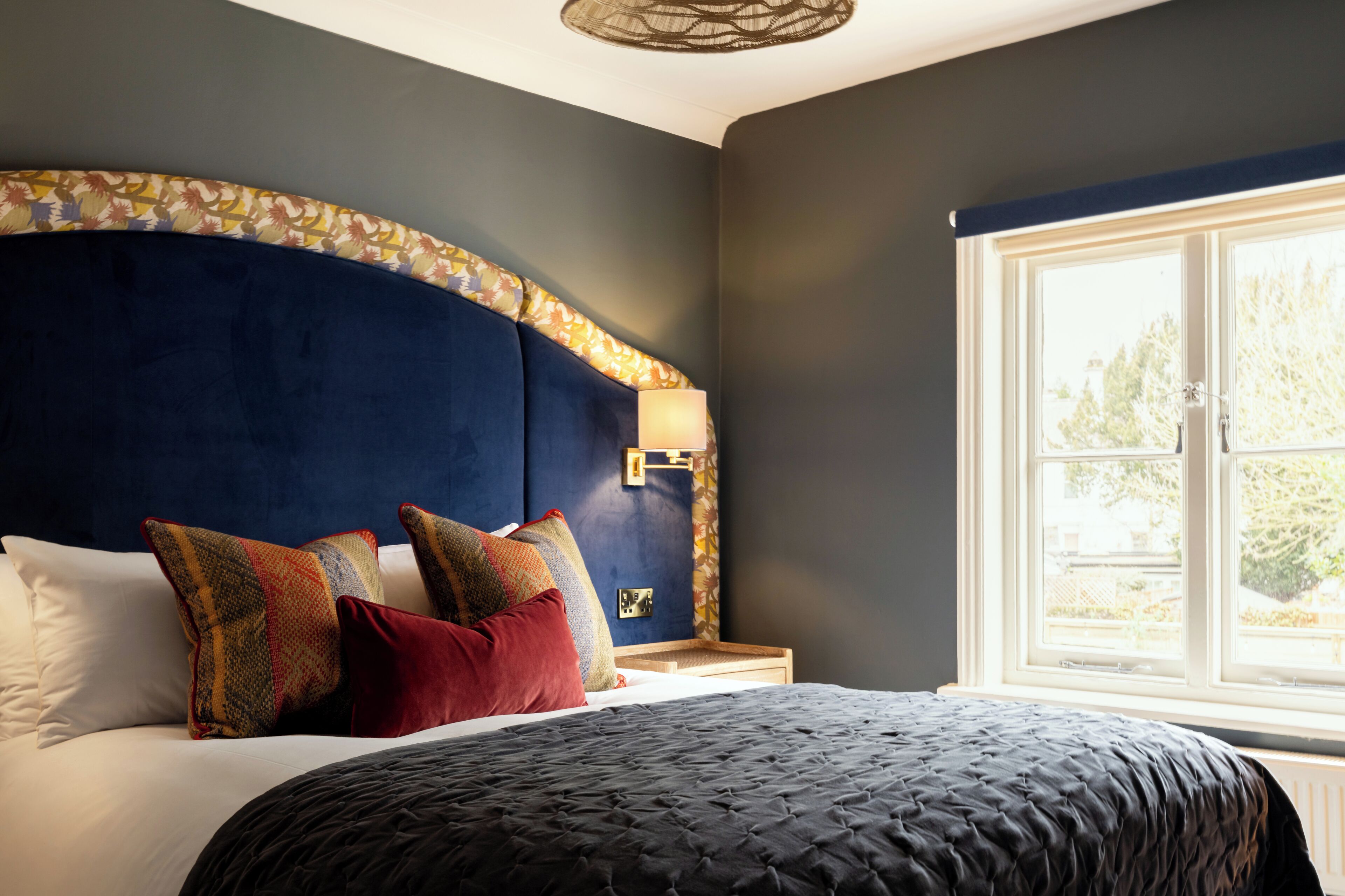 A bedroom with a large bed, a headboard with a floral pattern, a bedside table with a lamp, and a window with a view of greenery.