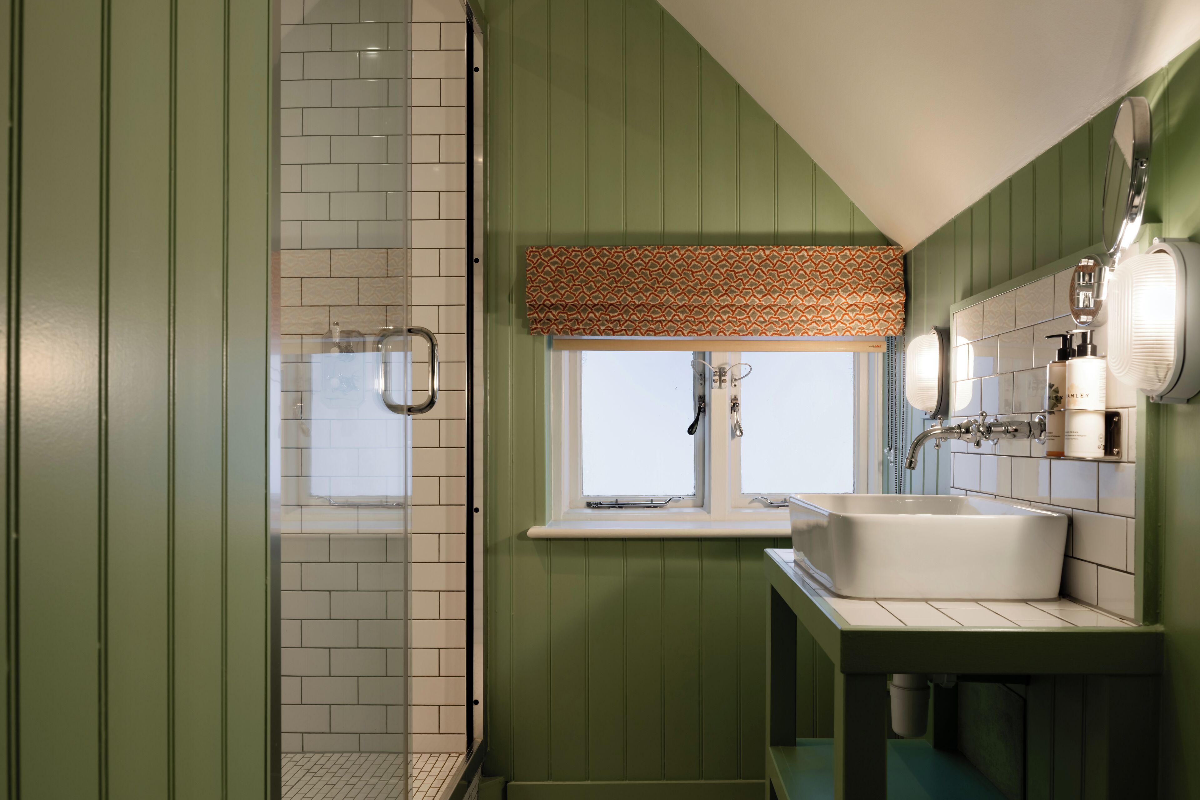 A bathroom with a shower, a sink, and a window with a patterned blind.