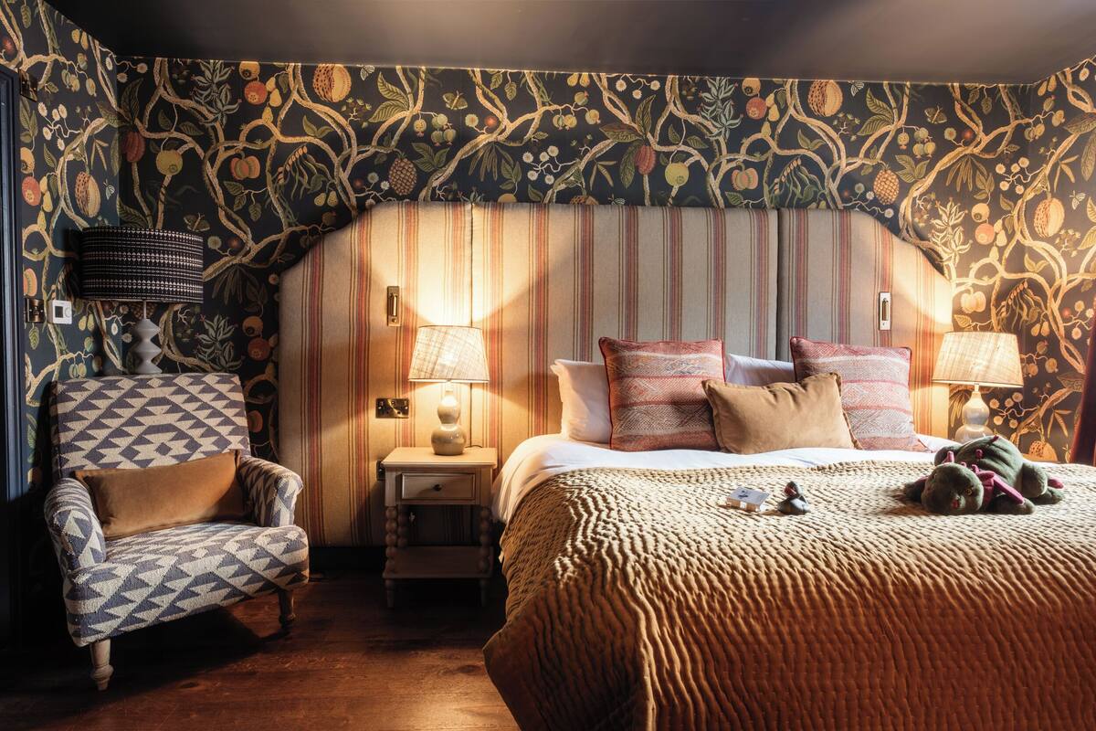 A bedroom with a patterned wallpaper, a striped headboard, a bed with multiple pillows, a chair, and two bedside tables with lamps.