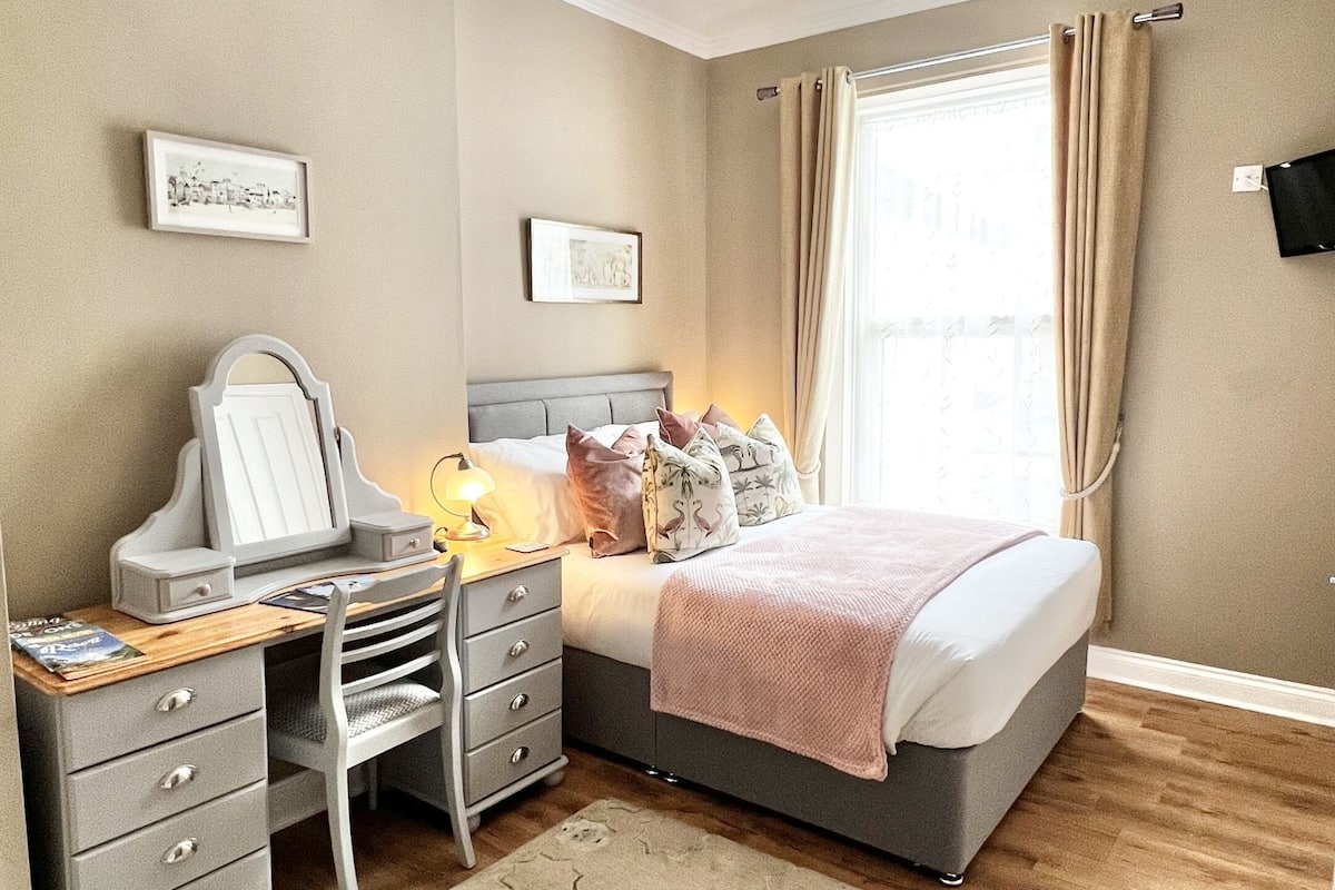 Un dormitorio con cama, escritorio con silla, espejo, lámpara y ventana con cortinas.