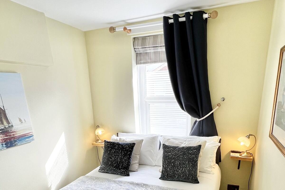 Un dormitorio ordenado con una cama, dos lámparas de noche, una ventana con cortinas y un cuadro en la pared.