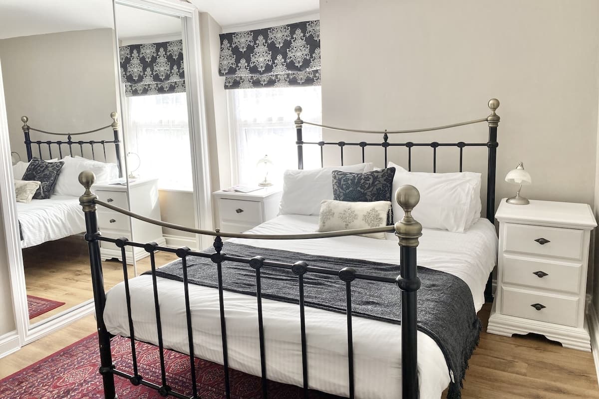Un dormitorio con cama con dosel, mesitas de noche blancas, un espejo y una ventana con cortinas estampadas.