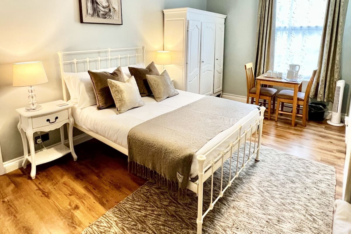 Un dormitorio con cama, mesitas de noche, lámpara, armario, mesa de comedor con sillas y ventana con cortinas.