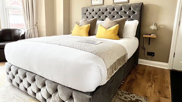 A bedroom with a tufted headboard, a large bed with white and yellow bedding, a patterned rug, and a nightstand with a lamp. - Devon House Guest House (Paignton)
