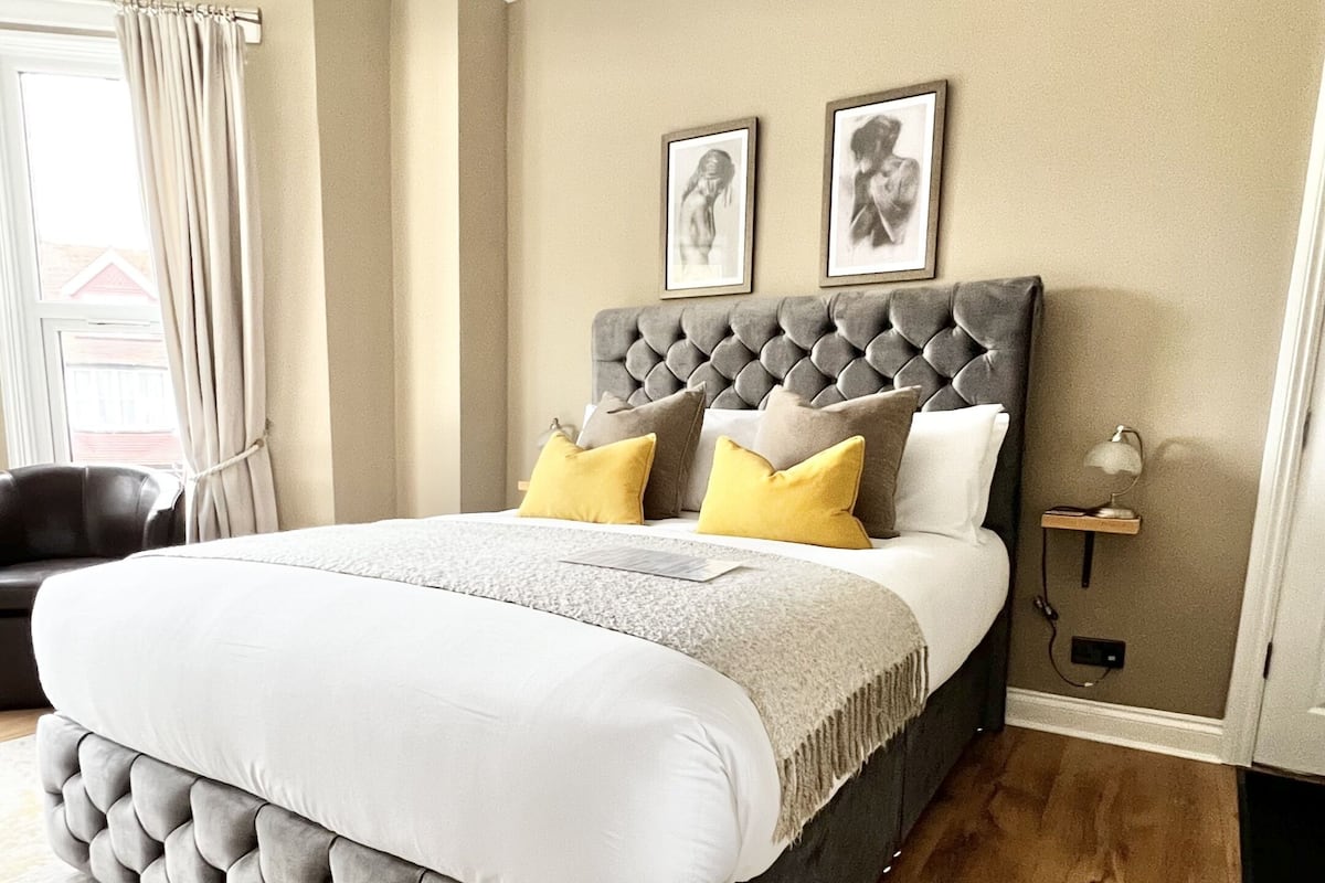 Un dormitorio con cabecera capitonada, cama grande con ropa de cama blanca y amarilla, alfombra estampada y mesita de noche con lámpara.